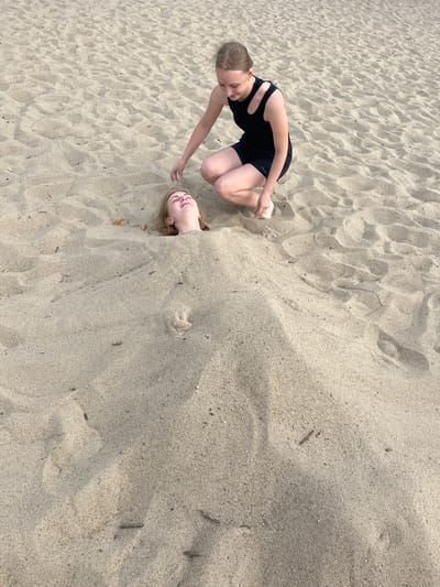 Two young women are buried in sand on a beach. The woman on the left is wearing a black swimsuit and is crouched down, reaching out with her hand. The woman on the right has her head and shoulders sticking out of the sand, with her eyes closed and a relaxed expression. The sand is light brown and textured, with some small twigs and debris scattered throughout. The lighting suggests it is daytime, possibly late afternoon, with shadows indicating the sun is at an angle. The overall mood appears playful and carefree. There is no visible text in the image.
