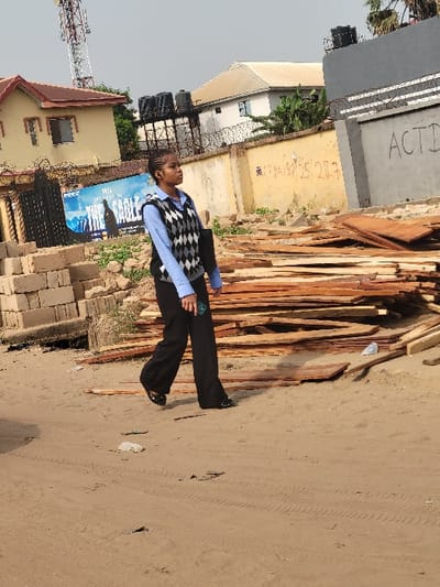 The scene depicts a young female walking rightward across a dusty, light brown dirt path in Puerto López, Colombia. The path exhibits tire tracks and minor debris, including white plastic fragments. The female, with braided hair, wears a light blue long-sleeved shirt, a black and white argyle V-neck sweater vest, dark trousers, and dark flat shoes. She carries a dark, rectangular object, possibly a folder or tablet, in her left hand.
To her left, a stack of light-colored concrete blocks rests near a dark metal gate. Behind these, a large billboard partially displays "THE EAGLE" and "2023." Further left, a two-story building with a reddish-brown roof and light yellow walls is visible, surmounted by a tall, red and white lattice telecom tower against a pale sky.
To her right and extending into the background, a substantial pile of rough-cut wooden planks lies on the ground. Beyond this, a low, light yellow wall topped with barbed wire runs horizontally. The wall has faint dark lettering and numbers visible, including "ACTD." Several black water tanks on metal frames are positioned behind this wall and on the roof of a more distant light gray building. Sparse green vegetation is discernible between structures.