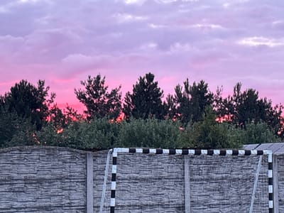 The image depicts a fence, a soccer goal, and a line of trees against a vibrant pink and purple sunset sky. The fence in the foreground is constructed of textured gray concrete blocks. To the right of the fence, a soccer goal with a white and black striped frame and a white net is partially visible. Behind the fence and goal, a dense line of various green trees forms a silhouette against the colorful sky. The sky itself is filled with soft, wispy clouds that are illuminated by the setting sun, creating a gradient of pink near the horizon transitioning to shades of purple and light blue higher up. There are no people or animals visible in the image. The scene appears to be an outdoor area, possibly a park or a backyard, located in the vicinity of Świba, Poland.