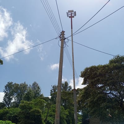 The image captures a street scene in Mweiga, Kenya. Two concrete utility poles stand prominently, one supporting a transformer and a tangle of electrical wires. A second, taller pole holds a cluster of floodlights at its apex. Overhead, multiple high-voltage lines stretch across the sky, intersecting the blue sky with scattered clouds. Lush green trees border the scene, their foliage filling the lower portion of the frame. A building is partially visible behind the tree in the lower right corner.