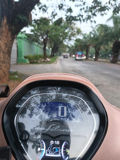 A photograph taken from the perspective of a stationary Yamaha Grand Filano scooter, located on a paved road in the Special Capital Region of Jakarta, Indonesia. The scooter's light brown dashboard occupies the foreground, displaying a digital screen with "0 km/h" indicating no movement and the time "6:18". Reflections of overhead trees and the overcast sky are visible on the dashboard's screen.
The road, appearing to be asphalt, extends into the midground and background. On the left side of the road, a substantial tree trunk is prominent in the immediate foreground, obscuring part of a tall, bright green wall with a darker upper border that runs parallel to the road. Beyond the wall, dense foliage is visible. On the right side of the road, a line of palm trees and other deciduous trees stands alongside the thoroughfare. In the background, two vehicles are present on the road: a blue and white vehicle, possibly a bus or large van, with illuminated headlights, followed by a smaller, dark-colored vehicle further behind it. The sky above is overcast, providing diffused lighting to the scene. No individuals are explicitly visible.