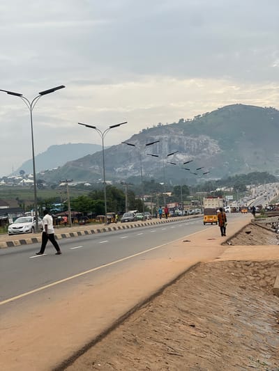 The image captures a wide-angle view of a busy road in Dam Camp, Nigeria, under an overcast sky. The foreground features a paved road with distinct white and black striped markings, and a yellow dividing line. A man in a white shirt and dark trousers is walking across the road, moving from left to right. The road is lined with streetlights on tall poles.
On the right side of the road, a dirt path runs alongside, bordered by a low stone wall. A man in a colorful patterned shirt and dark trousers is walking away from the camera on this path. Further down the road, there is a steady flow of traffic, including cars, minibuses, and a yellow auto-rickshaw.
In the background, the landscape is dominated by a large, rocky hill with patches of vegetation. A settlement of houses is visible on the lower slopes of the hill, and several utility poles with wires are present along the roadside. The overall scene depicts a typical urban environment with ongoing daily activities.