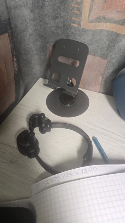 The image shows a close-up, slightly overhead view of a desk with a notebook, a phone stand, and a pair of novelty headphones. The notebook is open to a page with blue graph paper, and a handwritten mathematical equation is visible: "1) 32500/25 = 1300". The phone stand is black, with a circular base and a vertical arm that holds a phone in place. The headphones are black and designed to look like hands giving a thumbs-up. A blue pen rests on the notebook. The background is a textured fabric surface, possibly a sofa or curtain. The lighting is artificial and casts shadows. There is no discernible indication of the location in Ukhta, Russia.
