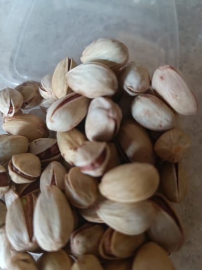 A close-up, vertical, medium shot captures a pile of pistachios inside a clear plastic container. The pistachios are light beige with some displaying a reddish-brown hue, and most are split open revealing the edible nut inside. The background is blurred, suggesting a shallow depth of field, and appears to be a light-colored, possibly textured surface. The lighting is even, with no strong shadows. The scene is static, with no discernible action or interaction taking place. The location in Kipshek, Russia, is not visually evident from the image.
