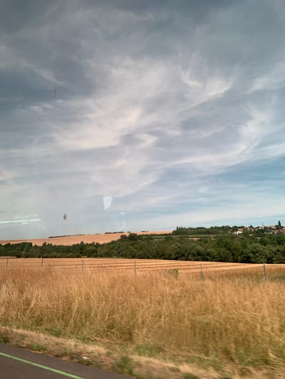 The image depicts a rural landscape with a field of golden wheat in the foreground, stretching towards a line of green trees and a village in the distance. The sky is filled with a mix of dark and light clouds. The scene appears to be captured from a moving vehicle, as indicated by the motion blur in the lower portion of the image and the reflection of what seems to be a window on the left side. The village, located in Moineville, France, is nestled amongst the trees on a gentle hill, with several houses and a church steeple visible. The overall impression is one of a serene, sun-drenched countryside.