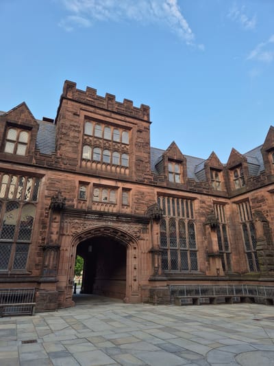 The photograph captures a view of a Gothic Revival building on a clear day in Princeton, New Jersey, United States of America. The building is constructed of reddish-brown stone, featuring crenellated towers, pointed arches, and numerous Gothic-style windows with diamond-shaped panes. A prominent arched entryway is visible in the center of the frame, leading into a dimly lit interior. The exterior is adorned with decorative stonework. In front of the building, a courtyard area is paved with large, irregular flagstones. A metal bench with a stone seat is positioned to the left of the entryway, and a low metal fence with a stone seating area runs along the front of the building to the right. The sky above is a clear blue with a few wispy white clouds. The overall scene suggests an architectural institution, possibly a university campus, given the style and grandeur of the structure. No people are clearly visible or engaged in any discernible activity within the frame.