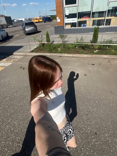 The image appears to be a selfie taken outdoors in Kazan, Russia. The perspective is from above, looking down. The subject is a person with reddish-brown hair wearing a white top and black-and-white patterned shorts. Their arm, which holds the camera, is visible in the lower right, showing a tattoo. The shadow of the person's hand is cast on the asphalt.
In the background, there is a building with large windows, landscaping including shrubs and flowers, and a parking area with several vehicles, including a grey car. Further back, there are buses. The sky is blue with some clouds.