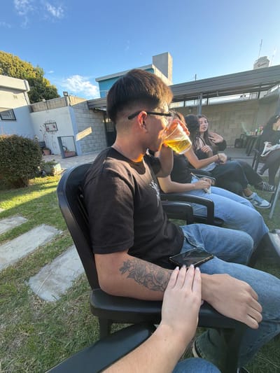 The scene depicts an outdoor social gathering in a residential backyard in Quilmes Oeste, Argentina, under a clear blue sky. In the foreground, a male individual is seated in a black plastic chair, shown in profile facing right. He has short, dark hair, wears dark-framed glasses, and a black short-sleeved t-shirt. He is drinking a light-yellow liquid from a clear plastic cup held in his right hand. A tattoo featuring roses is visible on his right forearm. A smartphone with a dark case and a cracked screen rests on his left thigh. A light-skinned hand with light pink painted fingernails is resting on his left forearm.
Behind the primary subject and to his right, a second person is partially visible, also seated in a black plastic chair. Further back, a female individual with long dark hair is seated in a black plastic chair, her right arm raised in a gesture. At least two more individuals are partially visible further to the right. The ground consists of a grassy lawn with concrete stepping stones. In the background, residential structures include a light-colored building with a window on the left, and a concrete block wall with a light blue section, topped by a dark metal pergola structure. A white water tank is visible on a distant rooftop.