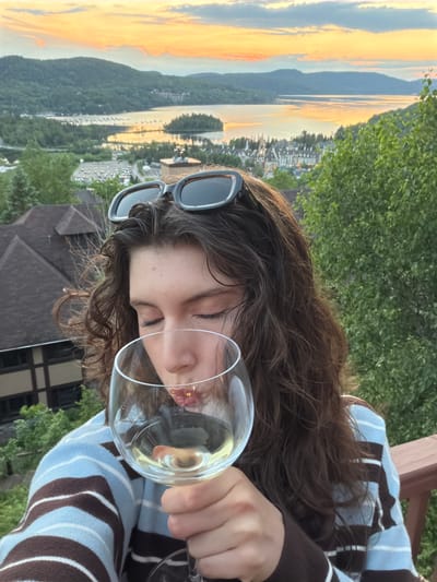 A young woman with long, curly brown hair and sunglasses perched on her head is holding a glass of white wine and drinking from it. She is wearing a blue and brown striped sweater. The background shows a scenic view of Mont-Tremblant, Quebec, Canada, with a lake, rolling hills covered in trees, and buildings in the distance, all bathed in the warm glow of sunset. The image appears to be taken from a balcony or deck, indicated by the presence of a wooden railing in the foreground.