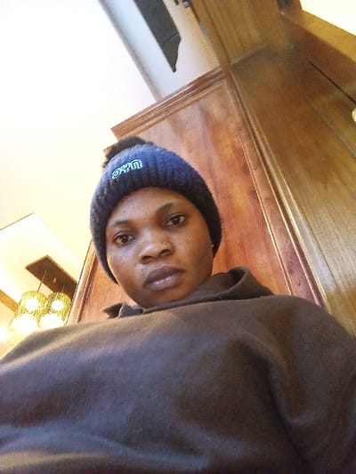 An individual is depicted in an indoor setting, likely a commercial establishment such as a cafe or restaurant, in an unspecified location within Madinaty, Egypt. The subject, a dark-skinned individual, appears to be female and is positioned in the upper center of the frame, looking directly into the camera. She wears a dark blue knitted beanie with a black pom-pom on top and light green embroidery, possibly reading "UAC" or "UGC," on the front. A small metallic stud, likely a nose ring, is visible on her left nostril. She is dressed in a dark, possibly black or dark brown, hooded sweatshirt.

The background to the right of the individual features vertical wooden paneling with a distinct decorative molding. To the left, a light-colored ceiling is visible, from which a light fixture hangs. This fixture consists of two cylindrical or basket-weave style shades, appearing to be made of a natural fiber, encased within a dark frame and emitting a warm, yellow light. The lighting suggests an artificial indoor environment. The perspective is from a low angle, looking upwards towards the subject and ceiling.
