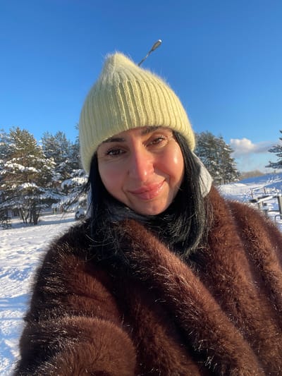 An outdoor portrait captures an adult female situated in a snowy environment within the city of Syasstroy, Russia, under a clear blue sky. The subject, positioned in the foreground, looks directly at the camera with a neutral expression and a slight smile. She wears a light yellow knitted beanie hat, a dark turtleneck or high-neck garment, and a brown fur coat, with a light grey hooded garment visible beneath the fur coat. Her dark hair frames her face, and her cheeks exhibit a rosy tone. The background reveals a landscape uniformly covered in white snow, with multiple snow-dusted evergreen trees. A black street light pole extends diagonally upwards from the upper center of the frame. On the far right, faint outlines of snow-covered outdoor seating structures, possibly benches or picnic tables, are discernible. A small, white cloud or vapor plume is visible on the distant horizon to the right. The scene is brightly illuminated by direct sunlight, indicating a clear winter day.