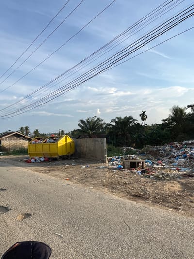 A bright yellow dumpster is overflowing with garbage. To the right of the dumpster, a large pile of trash, including plastic bags and debris, is scattered across the ground. In the background, palm trees and other vegetation are visible, along with some buildings and a gray concrete wall. Overhead, multiple electrical wires are strung across the sky, which is partly cloudy. The scene appears to be a roadside dump site in Alurcucur, Indonesia.