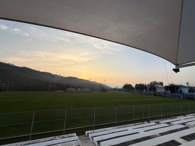 The image displays an outdoor football (soccer) training complex located in El Barrial, Mexico, at either sunrise or sunset, characterized by the warm, low-angle light and a sky transitioning from orange and yellow at the horizon to light blue overhead, with sparse cloud cover. The top portion of the frame is occupied by the underside of a large, light-colored fabric canopy or awning, with a support mechanism visible on the right. In the immediate foreground, a section of white concrete tiered bleachers is visible on the bottom right, separated from the playing area by a chain-link fence. The midground features multiple expansive, manicured green grass football fields, marked with white lines, extending into the distance. Several tall light poles equipped with floodlights are strategically placed around the fields. Beyond the fields, on the right side of the frame, several small, light-colored modular buildings are visible, alongside parked vehicles and additional structures. The background is dominated by a range of dark, tree-covered mountains. No individuals are present, and no activities are currently taking place on the fields.