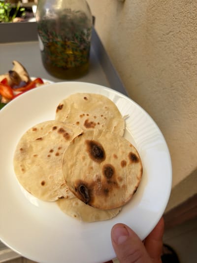 A close-up shot shows three flour tortillas on a white plate. The tortillas are lightly browned with some darker, charred spots. In the background, a glass bottle containing olive oil with herbs is visible, along with a blurred image of sliced vegetables and a portion of a plant. The scene appears to be outdoors, with a textured, light-brown wall occupying the right side of the frame. The photograph was taken in Even Yehuda, Israel.