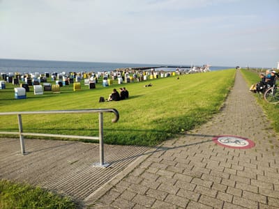 The image depicts a coastal scene in Cuxhaven, Germany, likely at theioxidizerdeich or a similar location along the Elbe estuary. The foreground features a paved walkway with a bicycle restriction sign, bordered by a grassy embankment and a metal railing. The embankment is dotted with numerous colorful beach chairs, some occupied by people. Beyond the embankment, the water of the North Sea stretches to the horizon under a clear sky. In the distance, a pier or dike is visible, with boats docked and people walking. The overall impression is one of a leisurely day at the seaside.
