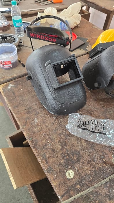 The image captures a collection of safety equipment and tools laid out on a worn wooden workbench, likely in a workshop or industrial setting in Gangapur, India. The scene is viewed from an overhead perspective, with objects arranged diagonally across the frame.
Prominently featured are two black welding helmets, one of which has a yellow hard hat resting on its top. Another piece of eye protection, possibly a face shield or safety glasses, has a black and red strap clearly labeled "WINDSOR" in red text. Adjacent to these items is a clear plastic bag with "HALLMARK SAFETYWEAR" printed on it.
Other objects scattered on the table include a clear plastic water bottle with a blue cap, which appears to be a "BISLERI" bottle. Nearby are several white work gloves, various hand tools such as wrenches and screwdrivers, and a metal disc that resembles a clutch plate or grinding wheel component. A clear plastic container with a red lid is also visible. The workbench itself is made of dark, scuffed wood, showing signs of heavy use with various marks and what appears to be dust or debris. A plank of raw wood extends from beneath the table on the right side. No people are visible, and no specific activity is taking place, though the arrangement of items suggests readiness for work or a temporary pause. The lighting indicates an indoor setting, likely during daytime.