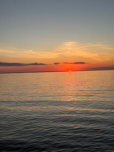 The image depicts a serene sunset over a body of water, likely the Kyiv Reservoir, near Vyshhorod, Ukraine. The sun, a vibrant orange orb, is low on the horizon, casting a warm glow across the sky and reflecting on the rippling water. The sky transitions from a pale blue at the top to shades of orange and pink closer to the horizon, with wispy clouds scattered throughout. The water is calm, with gentle waves creating a textured surface. There are no people or man-made structures visible in the frame, emphasizing the natural beauty of the scene. The overall impression is one of peace and tranquility.