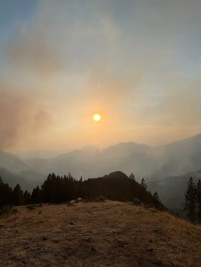 The image depicts a mountainous landscape at sunset, with the sun appearing as a bright, golden orb low in the hazy sky. The atmosphere is thick with smoke, creating a warm, orange-tinted haze that obscures the distant mountain ranges, giving them a layered, ethereal appearance. In the foreground, a dry, grassy ridge slopes downwards, dotted with rocks. A dense line of dark pine trees forms a sharp contrast against the smoky sky along the crest of the ridge. The scene suggests a remote, natural environment, potentially affected by wildfires given the smoke. No people or specific structures are visible. The location is not San Juan de Plan/San Chuan de Plan, Spain, as the landscape does not match the known geography of that area.