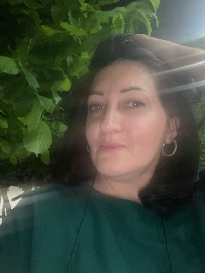 A woman poses for a selfie outside at night. She has dark hair, fair skin, and is wearing a green top and hoop earrings. She is touching her hair with one hand. Lush green foliage is visible in the foreground. The image was taken in Verin Ptghni, Armenia.