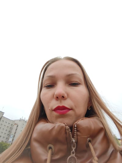 A close-up, low-angle selfie of a woman with long blonde hair, wearing a brown jacket. Her face is the primary focus, with bright red lipstick as a prominent feature. A faint mole is visible on her cheek. She is wearing a small gold earring in her visible ear. The background shows a cloudy sky and a multi-story building with many windows, suggesting an urban environment. The perspective gives the impression the photo was taken outdoors, possibly on a street or sidewalk. The overall mood appears neutral, with no strong emotional expression. The time of day is not clearly indicated, but the overcast sky suggests it might be daytime.