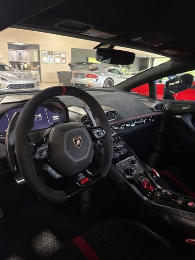 The image shows the interior of a Lamborghini, focusing on the steering wheel and dashboard. The steering wheel is black with red stitching and features the Lamborghini emblem. The dashboard includes various controls and a display screen. Outside, seen through the windshield and windows, are other vehicles and what appears to be a dealership or showroom setting. The location is potentially in West Chicago, United States.