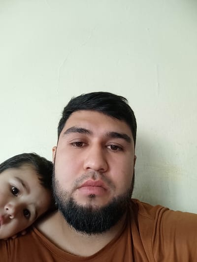 A selfie shows a man with dark hair, brown eyes, and a full black beard. He wears a brown t-shirt and has a serious expression. A young child with dark hair rests their head on the man's shoulder, looking directly at the camera with a curious expression. The background is a plain light-colored wall. The photo is taken at a close-up angle, focusing on the faces of the man and child.