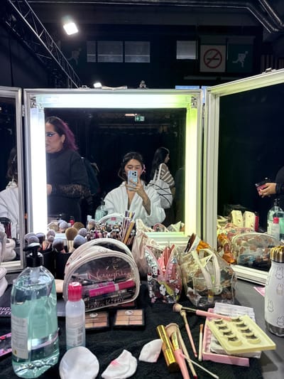 The image depicts a backstage or dressing room environment located in the city of Issy-les-Moulineaux, France. The scene is dominated by a three-panel vanity mirror setup featuring prominent fluorescent tube lights, one of which is visibly branded "OSRAM."

Reflected in the central mirror panel is a young woman with light brown hair, partially secured with pink clips, wearing a white bathrobe. She holds a light blue smartphone, actively capturing a selfie. To her reflected left, a person with purple hair, wearing a black long-sleeved top with lace detailing and dark eyeliner, is partially visible, looking towards the left of the frame. Further right in the reflection, another individual with dark hair, wearing a striped top, is seen from behind. A hand holding a dark container is also visible on the far right within the reflection.

The foreground consists of a black surface, likely a table covered with a cloth, densely populated with makeup tools and products. This includes numerous makeup brushes arranged in various containers and spread across the surface. Multiple clear cosmetic bags, some bearing "Hello Kitty" branding, are present, containing items such as lip liners and makeup pencils. Two makeup palettes are visible on the black cloth. Additional items include a large clear pump bottle of light blue liquid, a smaller white and pink bottle, several used cotton pads with red residue, a tray of false eyelashes, a gold spray bottle, and a white insulated bottle adorned with black Miffy rabbit motifs.

The background behind the mirrors reveals a dark, industrial space characterized by exposed metal trusses and overhead lighting fixtures. On the back wall, a green "exit" sign displaying a running person pictogram and a red "no smoking" sign with a cigarette pictogram are visible.