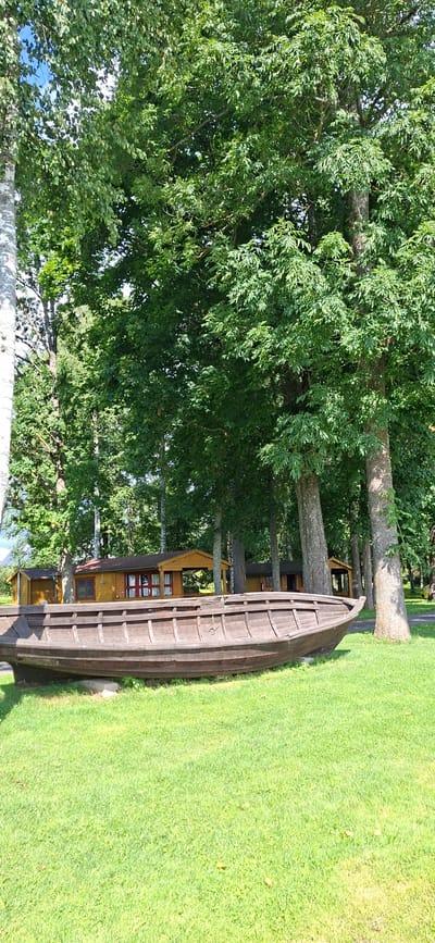 An outdoor, eye-level, vertical shot shows a weathered, wooden boat resting on a grassy lawn in front of a row of small, wooden cabins. The boat is positioned horizontally across the lower portion of the frame, its dark wood contrasting with the vibrant green grass. Behind the boat and grass, a line of single-story, light-brown cabins with red-trimmed windows and doors are nestled amongst tall, leafy green trees. The cabins appear to be part of a tourist accommodation or resort. The trees create a dense canopy overhead, with dappled sunlight filtering through the leaves. The sky is a bright blue with a few wispy clouds. The scene suggests a peaceful, natural setting, likely in Valma, Estonia, a town known for its natural beauty and potential for outdoor recreation. There are no people visible in the image.