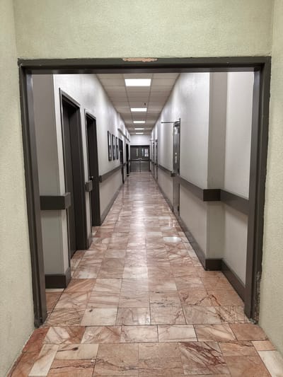 The image shows a long, empty hallway with a tiled floor. The hallway is viewed from an open doorway. The walls are painted a pale color, and there are doors on both sides of the hallway. The floor is tiled with a pattern of pinkish-brown and cream-colored tiles. There are fluorescent lights on the ceiling. The hallway extends into the distance, where it appears to turn a corner. The scene is well-lit and appears to be indoors. The location is in El Paso, United States of America.