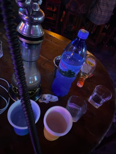 The image depicts a wooden table in a dimly lit setting with a hookah, various drinks, and empty glasses. The hookah is in the foreground, with its black hose and silver base. On the table, there is a blue plastic bottle of water labeled "Fiza Water," a clear glass bottle with a yellow label, and four drinking glasses - two white plastic cups filled with liquid, one clear glass filled with liquid, and one empty clear glass. The background is blurred and shows a few people out of focus. The image appears to have been taken in an establishment serving drinks and food, possibly a restaurant or bar. The location, based on the provided coordinates, is in the city of Dodoma, Tanzania.