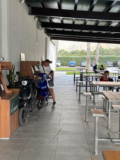 In Carini, Italy, two young men are lounging on the patio of an outdoor dining area. The man closer to the foreground is sitting on a black scooter, wearing a white t-shirt and white shorts. He has a bag slung over his shoulder. He is looking to his left, with his right leg extended. Behind him is a blue scooter.
The scene depicts an outdoor seating area with tables and benches, set against a backdrop of lush green foliage and distant hills. The architecture features dark wooden beams and grey tiled flooring, suggesting a modern and contemporary design. In the background, several cars are parked, and other patrons are seated at tables. The overall atmosphere appears relaxed and casual, possibly during daylight hours with pleasant weather. The image is oriented vertically, with the sky and foliage at the top.