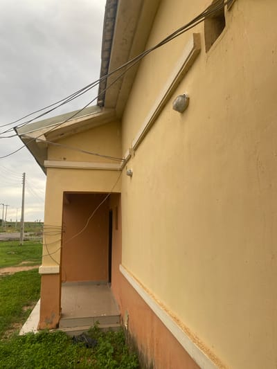 The image depicts the exterior of a building with a light yellow facade. A white molding runs along the edge of the overhang. There are two outdoor light fixtures mounted on the ceiling, and electrical wires are visible, hanging loosely. To the right, there is an entrance to the building, with a brown-colored door and a tiled walkway. The ground is covered with green grass and some dirt. In the background, there are utility poles with wires, indicating a semi-urban or rural setting. The sky is overcast, suggesting a cloudy day. The image is oriented sideways. There is no visible text.