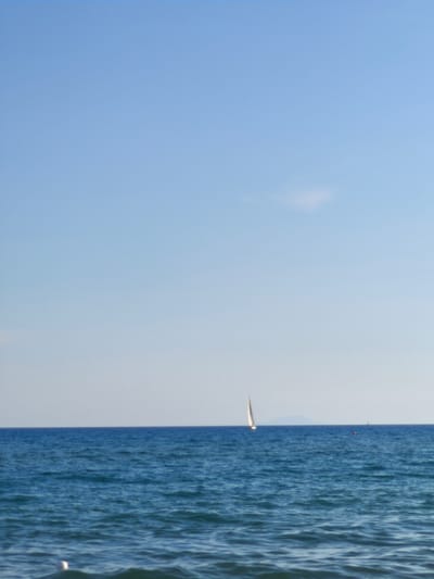 The media file depicts a serene seascape with a clear, pale blue sky dominating the upper portion of the image. A vast expanse of deep blue ocean fills the lower half, with gentle waves creating a textured surface. On the horizon, a small white sailboat with its sail fully extended is visible, appearing to be in motion. Further in the distance, another, smaller sailboat can be seen, and a red buoy marks a point in the water. A faint outline of a distant landmass, possibly an island, is visible on the horizon behind the sailboat. In the foreground, at the very bottom of the image, a white object, likely a buoy or part of a marker, partially submerged in the water, is visible. The lighting suggests it is daytime, likely morning or late afternoon, given the soft quality of the light and the absence of harsh shadows. The overall impression is one of tranquility and peacefulness. The location context provided indicates this scene is Castiglione della Pescaia, Italy. There is no visible text in the image.