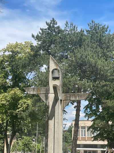 This is a vertical, eye-level, outdoor photograph taken in Kubrat, Bulgaria. The image depicts a modern concrete structure with geometric angles, possibly a monument or part of an architectural installation. A cylindrical object, possibly a speaker or light fixture, is visible within an opening in the concrete.

The structure is surrounded by lush green trees, including a large pine tree with its needles prominent in the upper portion of the image. The foliage is dense and appears to be in full bloom, suggesting a warm season. Behind the trees and structure, a building with a light-colored facade and multiple windows can be seen. The sky is blue with scattered white clouds, indicating fair weather.

The scene is static, with no discernible activity or people. The lighting suggests it is daytime, likely midday or afternoon, due to the bright sunlight casting shadows. The overall impression is one of an outdoor public space, possibly a park or plaza, in a built-up area given the presence of the building. No text is visible in the image.