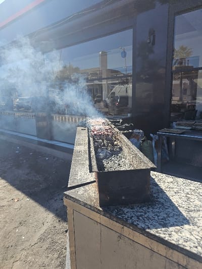 An outdoor commercial scene in Boufakrane, Morocco, features a long, rectangular charcoal grill actively cooking multiple metal skewers of meat, generating significant white smoke. The grill, containing hot coals and ash, rests on a speckled granite countertop. This setup is positioned directly in front of a building characterized by large, dark, reflective glass windows. The reflections in these windows reveal an urban street environment, including parked vehicles (a white van and several cars) and a clear blue sky. Stacks of white plates are discernible through the reflection on the right side of the building's interior. The ground immediately in front of the counter is unpaved dirt. The scene is illuminated by strong daylight, creating distinct shadows. No individuals are directly visible.