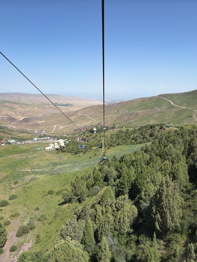 A wide-angle, high-angle shot captures a panoramic view of a mountainous landscape on a clear day, likely taken from a cable car in Kashka Suu, Kyrgyzstan. The sky is a vibrant, cloudless blue. A thick black cable, part of the cable car system, descends diagonally from the top left corner of the frame.

In the mid-ground, a cluster of small, white, yurt-like buildings forms a settlement nestled in a verdant valley. A winding dirt road snakes through the landscape, connecting various points. The slopes of the surrounding hills are covered in green grass and scattered trees, with patches of bare, dry earth visible in some areas.

Closer to the foreground, a dense forest of evergreen trees, primarily junipers, dominates the scene. The trees vary in height and density, creating a textured carpet of green. Some areas of the hillside show lighter green vegetation, possibly fields or meadows.

On the right side of the frame, a section of the cable car line is visible with several empty seats suspended in the air. The cable car seats are colored blue and yellow. The overall impression is one of natural beauty and a rural, mountainous environment.