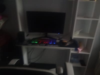 The image shows a dimly lit room, likely a bedroom or a home office. The main subject is a computer setup on a desk, which includes a monitor, a keyboard with RGB lighting, and a speaker. To the right of the desk is a white shelving unit with several compartments, some of which are empty and some contain various items that are not clearly discernible due to the low resolution. The overall scene suggests a personal space, possibly used for gaming or work. The lighting is low, indicating it might be nighttime or the lights are intentionally dimmed. There are no people visible in the image, and no specific activity is clearly depicted beyond the presence of the computer equipment. The location cues are limited to the general interior setting.
