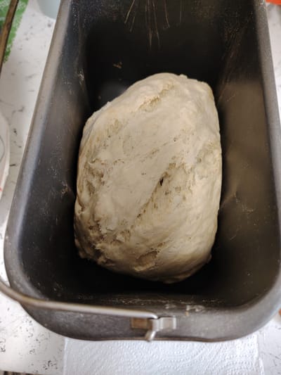 A close-up, overhead shot shows a light-colored, uncooked dough ball resting inside a dark gray bread pan. The dough is lumpy and textured, with visible indentations and air pockets. The bread pan has a slight sheen and appears to be made of metal. The background is blurred but suggests a white, speckled surface and a white paper napkin. The image does not contain any identifiable elements that would allow for the determination of its location in Vladimirovka, Russia.