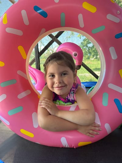 A young girl with brown hair, wearing a colorful swimsuit, smiles as she lies on her side within a large, pink inflatable donut. The donut has white, yellow, blue, and green sprinkles. Behind the girl, a pink inflatable flamingo can be seen. The background shows greenery and a dark, possibly netted structure, suggesting an outdoor setting. The girl's arms are crossed over her chest, and her hands are visible. The lighting indicates it is daytime. The girl appears happy and relaxed.