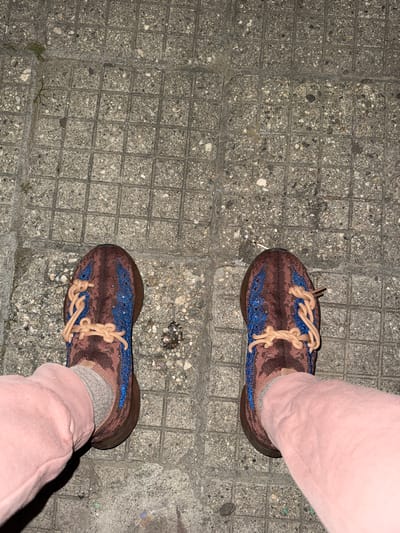 The image shows a person's feet wearing brown and blue sneakers with light brown laces and light pink sweatpants, standing on a sidewalk composed of square concrete blocks. The sidewalk appears to be slightly weathered and stained in some areas. The picture has been taken from an elevated perspective, looking down at the feet. The scene does not provide enough details to discern the specific location within the city of Polena, Bulgaria. No interactions or actions are visibly occurring.