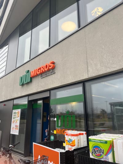 The image captures the exterior of a Migros supermarket, presented at an oblique angle with a counter-clockwise rotation, making the building appear diagonal. The modern facade features large glass windows and a light-colored, concrete-like material.

A prominent store sign with orange lettering spelling "MIGROS" is displayed, accompanied by a green, stylized 'M' or leaf-like symbol below it. Smaller, less distinct text below "MIGROS" appears to read "PARTENAIRE."

Outside the store's entrance, stacks of paper towels or kitchen rolls are visible on black plastic crates. Brands identified include "Plenty" (with "Kompostierbare Tücher," meaning "Compostable Wipes," visible on its green packaging) and "TWIST" (on white packaging with "Action" and "Arbre" discernible).

The large glass windows reflect the surrounding environment, showing trees and what appears to be an overcast daytime sky. Interior lights, including a large circular fixture and another with a yellow, possibly decorative, pattern, are also reflected. Through the glass, a person can be partially seen near the entrance, suggesting activity. The green Migros logo is clearly reflected on the glass door.

A poster advertising food items, displaying fruits, vegetables, and bread, is visible through a lower window, with the word "AKTIONEN" (Promotions) readable. Part of a bicycle, including handlebars and a front wheel, is visible in the lower right corner. The presence of Migros, a major Swiss retail chain, aligns with the Meyrin, Switzerland context.