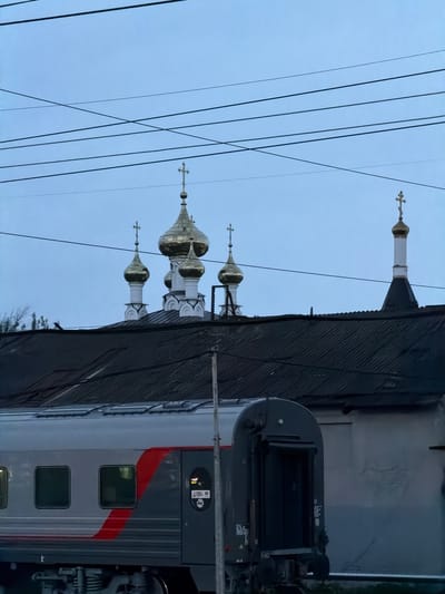 The image depicts a scene in Rtishchevo, Russia, juxtaposing a railway environment with the distinctive architecture of Russian Orthodox churches. In the foreground, a passenger train car is visible, marked with a red stripe, indicating that this is a train in Russia. In the background, the golden domes of a church are visible, towering above the train. Power lines intersect the view, spanning the sky between the train and the church domes. The photograph is taken in the evening, given the low light and the dark clouds.