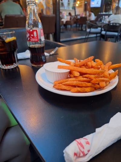 The image depicts a close-up view of a dining table in an outdoor or semi-enclosed restaurant setting in Petrich, Bulgaria, during what appears to be late afternoon or early evening. In the foreground, a white oval plate holds a serving of orange sweet potato fries alongside a small white cup containing a light-colored dipping sauce. To the left of the plate, a classic glass bottle of Coca-Cola with a red label is positioned upright. Further left, a clear glass contains a dark beverage, likely Coca-Cola, with ice and a lemon wedge. A crumpled white paper napkin with red markings is visible in the bottom right corner of the table. The table surface is dark, likely a laminate or textured metal.

The background is intentionally blurred, showing other patrons seated at tables. Several individuals are visible, including a person in a green shirt facing away from the camera, and another person with blonde hair further in the distance. The background also reveals various light sources, including warm indoor lighting, and what appears to be a television screen. The overall environment suggests a casual dining establishment with a moderate number of customers.
