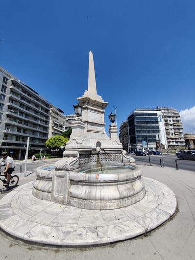 The image depicts a marble fountain in the center of a city intersection, with an obelisk rising from its center. The fountain features intricate carvings and is surrounded by a circular marble base. A cyclist is visible in the foreground, riding on the paved road to the left of the fountain. In the background, several multi-story buildings line the streets, with cars and pedestrians present. The sky is clear and blue. This fountain is located in Thessaloniki, Greece, and is known as the Fountain of the Obelisk or the Monument to the Conqueror.