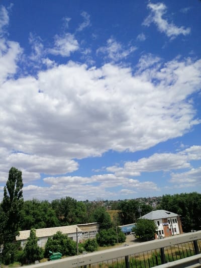 The image shows a view of a landscape on a cloudy day. In the foreground, there is a metal railing, suggesting the photo was taken from an elevated position like a bridge or balcony. Below the railing, there is a grassy area with some scattered vegetation.
In the middle ground, a long, low building with a corrugated metal roof stretches across the frame. It appears to be an industrial or storage facility, with a series of connected structures. To the left of this building, a large, mature tree with dense foliage stands prominently. A bright green dumpster is visible near the building. Further to the right, a modern building with a grey roof and brick accents is situated among the trees. It has multiple windows and appears to be a two-story structure. A blue semi-trailer truck is parked to the left of this building.
The background is dominated by a thick band of lush green trees, creating a natural border to the built environment. Beyond the trees, a residential area with houses and buildings can be seen on a gentle slope, leading up to the horizon. The sky above is a bright blue, punctuated by numerous white, fluffy cumulus clouds.
This scene appears to be located in Cernavoda, Romania, given the context provided. The overall impression is one of a partly industrial, partly residential area bordering a natural green space, under a dynamic sky.