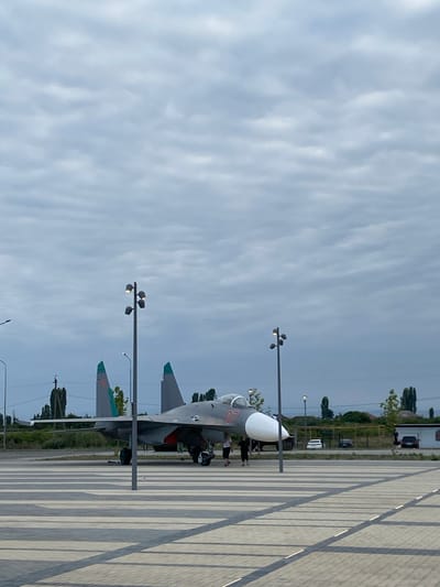 In Arablyar, Russia, a parked military jet with the number "07" visible on its nose cone is displayed on a paved area. The jet is gray with green accents on its tail fins and a white nose cone. Two streetlights are positioned in the foreground, casting shadows on the patterned paving stones. A few people are walking near the jet. In the background, there are trees, buildings, and several parked cars. The sky is overcast with gray clouds.