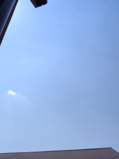 A low-angle, eye-level shot captures a clear, pale blue sky with a single, small white cloud on the left side of the frame. A dark, angled structure, possibly a building overhang or gutter, occupies the upper left corner of the image. The bottom of the frame is dominated by the top edge of a dark, corrugated roof, likely made of metal, extending horizontally across the scene. The scene is devoid of people or any visible activity, presenting a quiet, outdoor environment. The overall impression is one of a bright, possibly sunny day with a view upwards towards the sky and the edge of a building.