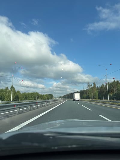 The image depicts a view from a vehicle traveling on a multi-lane highway in Protopopovka, Russia. The highway is asphalt with white lane markings. On the left, there is a metal guardrail with red reflectors. A dense line of green trees forms the background along the highway. Above the highway, power lines are strung, and several red and white spherical markers are attached to these lines. The sky is blue with scattered white clouds. A white truck is visible on the highway, moving away from the camera. The foreground shows the dashboard of the vehicle.