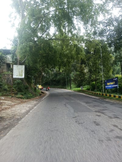 A winding asphalt road is depicted cutting through a densely forested, hilly environment in the city of Sat Thay (East), Myanmar. The road, which shows patches of repair and dampness, curves sharply to the left in the mid-ground. On the left side of the road, a dirt shoulder leads to a stone retaining wall, above which a partially obscured, light-colored building is visible amidst thick foliage. A large, faded white square sign with illegible text is mounted near the retaining wall. On the right side of the road, a black and yellow striped guardrail runs alongside. Further down the road on the right, a blue banner sign displays white text reading "Hati-Hati !!" and "Daerah Rawan Kecelakaan". Overhead, a thick canopy of green trees, some with drooping branches, covers much of the sky, indicating overcast conditions or a heavy tree cover. Three motorbikes are observed on the curved section of the road: two red scooters in the lead, followed by a yellow scooter or small three-wheeled vehicle further back. A street light pole is also visible on the right side of the road. No specific individuals are discernible on the motorbikes.