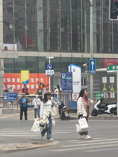 Here is a forensic description of the image:

The image shows a street scene in what appears to be Shenhe District, China. Several pedestrians are crossing the street. One woman, wearing a white jacket and jeans, carries a white shopping bag and a tan-colored bag. Another woman, wearing a plaid shirt and white jacket, also carries a white shopping bag. A man in a blue shirt and dark jacket is seen walking along the sidewalk.

The street is lined with signs, including one for "石化易电 Recharge" (Sinopec Easy Electricity Recharge) with parking fees noted. Other signs advertise development and productivity. There is a blue sign with a white arrow and Chinese characters indicating the direction for "机动车" (motor vehicles). A green and white sign indicates "惠宾 Huibin".

A multi-story building with a glass facade is on the left side of the frame, reflecting the street scene. Traffic lights are visible at the top left corner. Several parked scooters and motorcycles are also visible along the sidewalk. The setting seems to be an urban area.